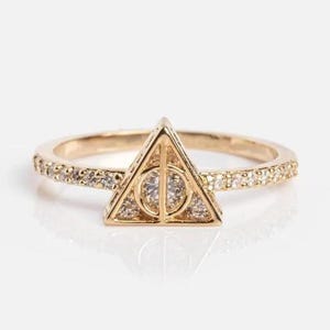 Może przedstawiać: Złoty pierścionek z symbolem Insygniów Śmierci. Pierścionek ma trójkątny element centralny z okręgiem i linią, a obręcz jest wysadzana małymi, przezroczystymi kamieniami. Pierścionek jest na białym tle.