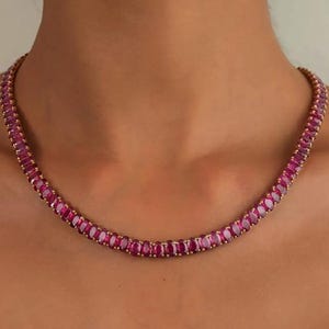 Könnte beinhalten: Eine goldene Halskette mit einer Reihe von oval geschliffenen, tiefroten Edelsteinen. Die Edelsteine sind eng aneinander gesetzt und bilden eine durchgehende Farblinie. Die Halskette wird am Hals einer Person präsentiert und zeigt ihr Design und ihre Farbe.