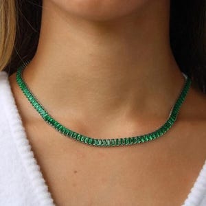 Puede incluir: Un delicado collar de color verde esmeralda con una cadena fina y pequeñas piedras rectangulares. El collar lo lleva una persona con una camisa blanca.