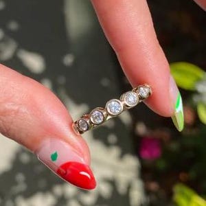 Könnte beinhalten: Ein goldener Bandring mit sieben runden, klaren Edelsteinen. Die Edelsteine sind in einzelnen Fassungen gefasst. Der Ring wird zwischen zwei Fingern mit lackierten Nägeln gehalten.