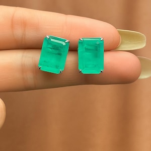 Puede incluir: Un par de pendientes de botón con piedras preciosas verdes esmeralda. Cada pendiente está engastado en un metal plateado con cuatro garras. Las piedras preciosas tienen forma rectangular y se muestran sobre un fondo neutro.