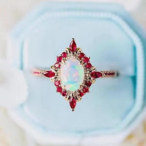 Könnte beinhalten: Ein goldener Ring mit einem ovalen, schillernden Opal-Mittelstein, umgeben von kleinen, roten Rubin-Akzenten. Der Ring wird in einer hellblauen Samtbox präsentiert, die das Design und den Farbkontrast hervorhebt. Der Ring hat ein feines, filigranes Band.
