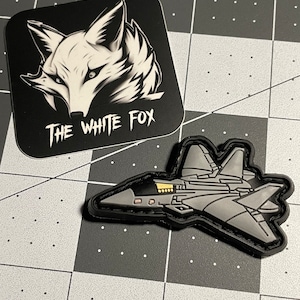May include: A black and white sticker with a white fox head and the text "THE WHITE FOX" next to a gray jet fighter patch with a black border. The items are on a black and gray grid.