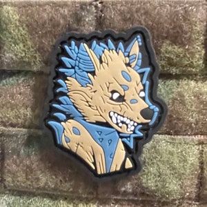 May include: A tan and blue cartoon wolf patch with a black border. The wolf has sharp teeth and is set against a camouflage background. The patch is likely made of rubber or a similar material.