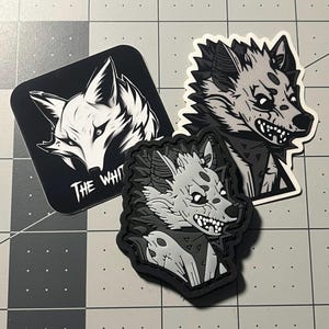 May include: Three black and white patches featuring stylized illustrations of wolves. The patches are on a gray grid background. The text "The White" is visible on one of the patches.