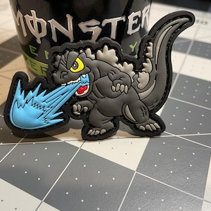 May include: A Godzilla patch in shades of gray, black, and white, with a blue flame effect emanating from its mouth. The patch has a black border and a yellow eye. The patch is on a gray and white surface.