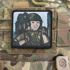 May include: A black and white embroidered patch featuring a cartoon illustration of a soldier wearing a helmet and holding a rifle. The soldier is wearing a green and brown camouflage uniform. The patch is rectangular and has a black border.