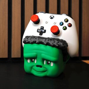 May include: A green figurine of a cartoon character's head with black hair, wearing a white Xbox controller. The controller has red and multi-colored buttons. The figurine has a sad expression.