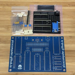 May include: A blue printed circuit board with the text picocomputer.github.io, accompanied by a plastic bag containing electronic components like resistors and pins, and a black foam block holding integrated circuit sockets and a blue VGA port.