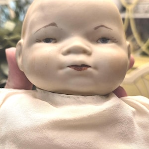 May include: A vintage porcelain baby doll with a serene expression. The doll has a pale complexion, blue eyes, and delicately painted eyebrows and lips. It is dressed in a cream-colored fabric.