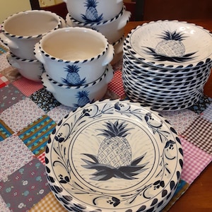 May include: A set of white ceramic bowls and plates featuring a blue and white pineapple design. The bowls have two handles and the plates have a scalloped edge.