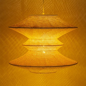 Op de afbeelding: Een drielaagse, gouden hanglamp gemaakt van geweven kralen. De lamp hangt aan het plafond en is verlicht.