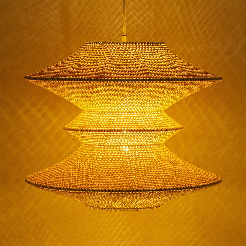Op de afbeelding: Een drielaagse, gouden hanglamp gemaakt van geweven kralen. De lamp hangt aan het plafond en is verlicht.