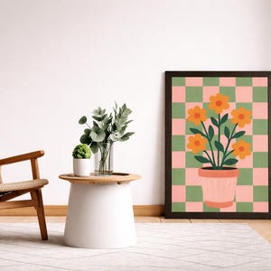 May include: A framed art print featuring a floral design with orange flowers in a pink pot against a green and pink checkered background. The artwork is displayed in a room with a chair, a small table, and decorative plants.