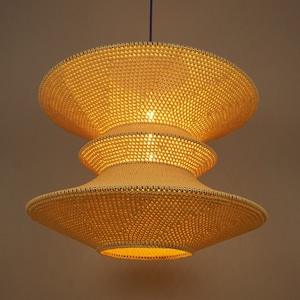 Op de afbeelding: Een drielaags geweven hanglamp met een warme gele gloed. De lamp is gemaakt van een natuurlijk materiaal, mogelijk rotan of bamboe, en heeft een uniek, modern ontwerp.