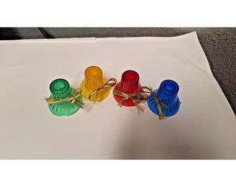 Könnte beinhalten: Vier farbige Kerzenhalter in Grün, Gelb, Rot und Blau. Jeder Halter hat ein goldenes Band um sich gebunden. Die Kerzenhalter sind aus einem durchscheinenden Material.