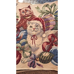 Könnte beinhalten: Ein Wandteppich mit zwei Katzen inmitten von Weihnachtsschmuck und Schleifen. Eine Katze trägt einen Weihnachtsmütze und eine rote Schleife. Die andere Katze ist braun mit blauen Augen. Der Hintergrund umfasst grüne, blaue und violette Ornamente.