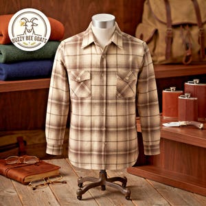 May include: A long-sleeved button-down shirt featuring a tan and brown plaid pattern with two chest pockets. The shirt is displayed on a mannequin torso. A circular logo in the upper left corner displays a goat illustration and the text Buzzy Bee Goats.