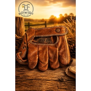 May include: A weathered brown leather baseball glove featuring a prominent wrist strap with a metal snap button closure. The glove displays visible stitching, a laced side panel, and a worn texture across the surface. The logo Buzzy Bee Goats is visible.