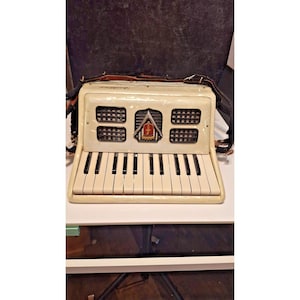 May include: Cream-colored accordion with black and white keys. The front panel features a decorative emblem and rectangular speaker vents. Leather straps are attached for carrying. The instrument is set against a dark background.