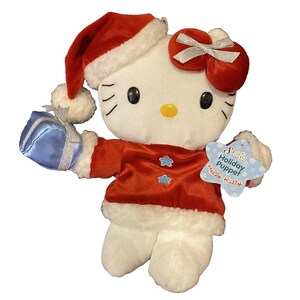以下が含まれることがあります： クリスマスの衣装を着たハローキティのぬいぐるみ。赤いサンタ帽と白いトリムのジャケットを着て、青いプレゼントを持っています。頭には銀色のリボンが付いています。「Sanrio Holiday Puppet Hello Kitty」と書かれたタグが付いています。