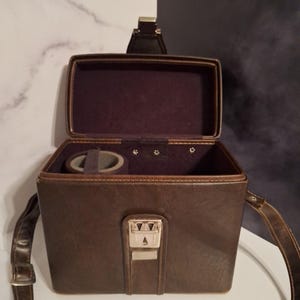 May include: Open brown leatherette vintage travel case with a handle and shoulder strap. The interior is lined with a dark purple fabric. The case has a silver metal clasp and a small compartment for accessories.