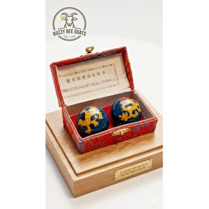 May include: A red, open box with two blue and gold health balls, resting on a wooden base. The box interior is red, and a paper with text is visible inside. The box is labeled "DIRECTIONOFHEALTH BALL."