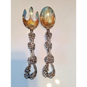 May include: A silver-toned serving fork and spoon set. The fork has three tines, and the spoon has an oval bowl. Both utensils feature handles adorned with grape and leaf designs, adding a touch of elegance to the serving set.