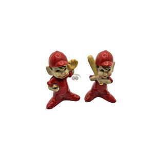 May include: Two ceramic baseball player figurines. Each figurine is wearing a red baseball uniform and cap. One figurine is holding a baseball and the other is holding a baseball bat. The figurines have elf-like features.