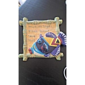 May include: A ceramic sign featuring a vibrant fish design and the words "Hawaiian Style: Please Remove Your Shoes." The sign is framed with a bamboo-style border and uses blue, yellow, and purple hues.