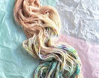 May include: A skein of hand-dyed yarn with a gradient of colors, from light beige to a speckled mix of blues, greens, and yellows. The yarn is displayed on a backdrop of crumpled tissue paper in shades of mint green, white, and pink.