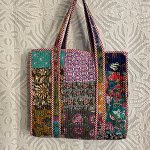 Puede incluir: Una bolsa de mano de patchwork con una variedad de patrones florales en tonos de azul, verde, morado y rojo. La bolsa tiene un ribete a rayas rojas y blancas y una sola asa superior.