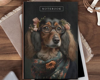 Könnte beinhalten: Ein Notizbuch mit einem Porträt eines Dackels mit Brille und einem Blumenschal. Das Wort "NOTEBOOK" ist oben aufgedruckt. Das Notizbuch liegt auf einem weißen Tablet, mit einem Stift und anderen Gegenständen im Hintergrund.