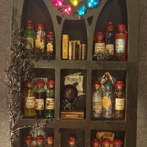 Puede incluir: Un gabinete de madera negro con múltiples estantes llenos de pequeñas botellas de vidrio con tapones de corcho y etiquetas. Las botellas contienen líquidos coloridos y están dispuestas en una cuadrícula. El gabinete tiene un diseño de arco gótico y está decorado con una vid de musgo.