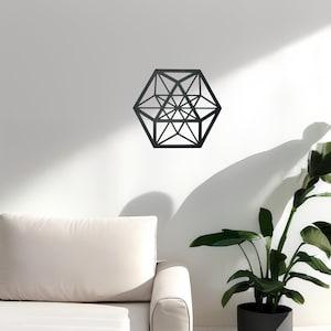 Pu&ograve; includere: Un decoro da parete geometrico nero a forma di esagono con effetto 3D. Il decoro &egrave; appeso a una parete bianca in un soggiorno con un divano bianco e una pianta in vaso.