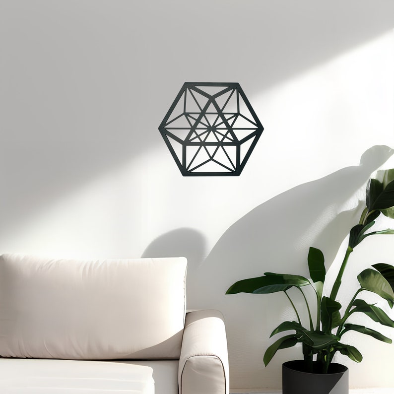 Pu&ograve; includere: Un decoro da parete geometrico nero a forma di esagono con effetto 3D. Il decoro &egrave; appeso a una parete bianca in un soggiorno con un divano bianco e una pianta in vaso.