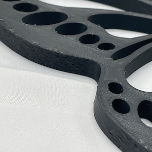 May include: A black plastic butterfly-shaped wall decor with a detailed design and multiple cut-out circles. The decor has a textured surface and a slightly curved shape.