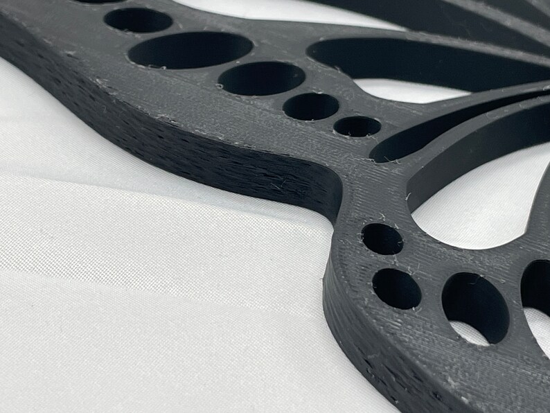 May include: A black plastic butterfly-shaped wall decor with a detailed design and multiple cut-out circles. The decor has a textured surface and a slightly curved shape.