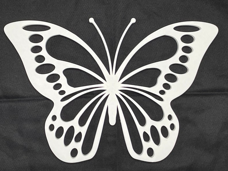 May include: White cutout butterfly wall decor with intricate details. The butterfly has a symmetrical design with a large body and wings with a pattern of circles and lines.