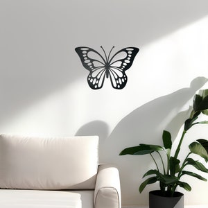 May include: Black metal butterfly wall decor with intricate details, mounted on a white wall with a white couch and a potted plant in the foreground.