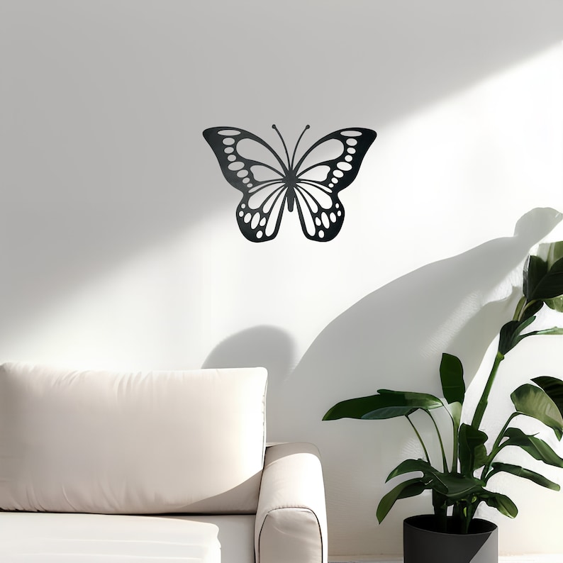 May include: Black metal butterfly wall decor with intricate details, mounted on a white wall with a white couch and a potted plant in the foreground.