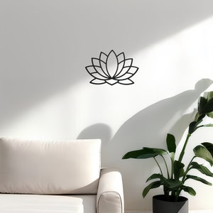 May include: A black metal lotus flower wall decor with a simple, geometric design. The decor is mounted on a white wall above a white couch and a potted green plant.