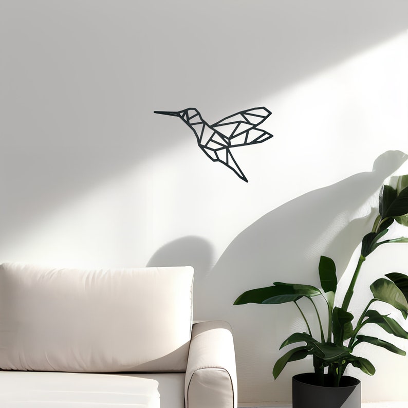 May include: A geometric black hummingbird wall decor hanging on a white wall above a white couch and a green plant in a black pot.