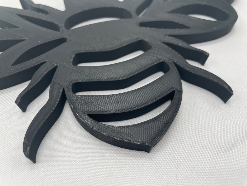 May include: A black 3D printed bee cutout with a detailed design. The bee has a segmented body and wings, and the wings are cut out to create a decorative effect.