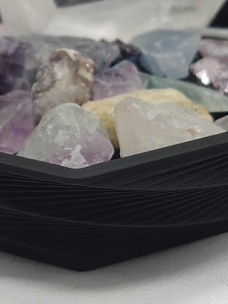 May include: A close-up of a black hexagonal tray filled with various colorful gemstones. The tray is made of a woven material and has a smooth, polished finish.