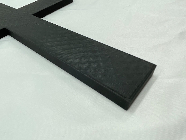 May include: A black cross made of a textured material. The cross is lying on a white surface.