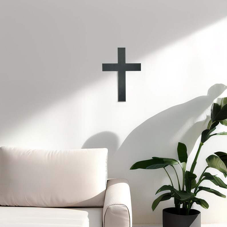 May include: A black metal cross mounted on a white wall. The cross is in the center of the image and is surrounded by a white wall. There is a couch and a plant in the background.
