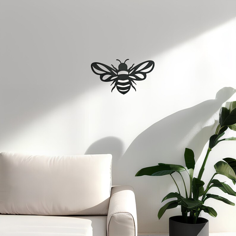 May include: Black metal bee wall decor with a simple design. The bee is shown in profile with wings spread out. The bee is mounted on a white wall with a white couch and a green plant in a black pot in the foreground.