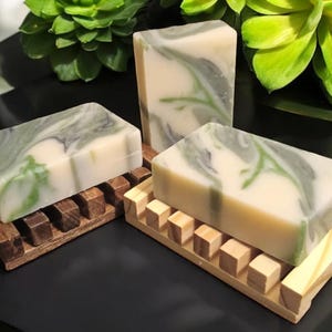 May include: Three rectangular bars of soap with a marbled green and white design, resting on wooden soap dishes. The soap dishes are made of dark and light wood. Green foliage is in the background.