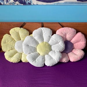 Könnte beinhalten: Drei plüschige Kissen in Blütenform in Gelb, Weiß und Rosa. Die Kissen liegen auf einem violetten Bettbezug.